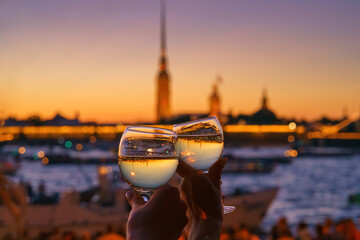 close-up view of two hands clinking glasses of champagne, Neva River and Peter and Paul Fortress are reflected upside down in glass, which are blurred in background, at evening, St Petersburg, Russia