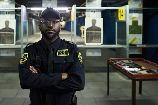 Portrait of African American security guard standing confidently with arms crossed at indoor shooting range. Targets and firearm training equipment visible in background