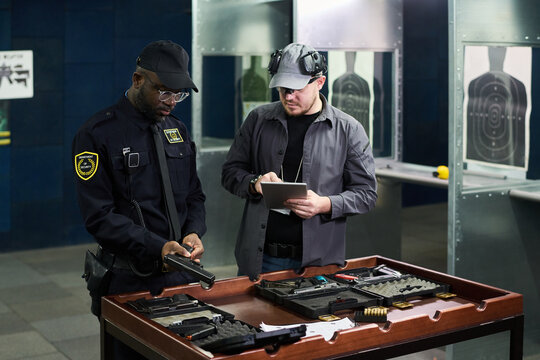 Police officer examining handgun while instructor taking notes in indoor shooting range training facility emphasizing safety protocols and gun handling techniques