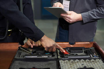 Individuals examining an array of firearms and equipment while using tablet for inventory. Detailed procedure involves pointing at various items laid out in organized manner