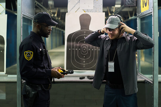 Two men standing at indoor gun range, discussing shooting results. One man, in security uniform, holding a flashlight, while other man adjusting headphones in front of target sheet