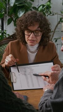 Medium vertical shot of confident young Caucasian female financial adviser explaining loan terms to mature couple during meeting in cafe, showing loan amortization schedule with monthly payments