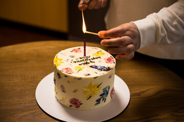 A hand is lighting a candle on a cake decorated with lovely floral designs, ready to enjoy. A hand...