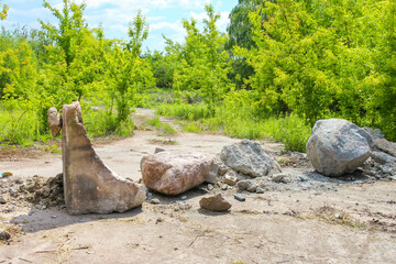 Two large, weathered concrete blocks sit on light brown earth with scattered rocks and green plants, against a blurred background of lush green trees, highlighting abandoned structures in nature,