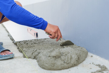 A worker hand uses a trowel to smooth gray mortar, preparing a level surface for floor tiles....