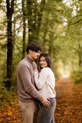 Fototapeta premium Side view of a young couple smiling and embracing in a forest during autumn. They look at each other with joy. Casual clothing. Concepts like love, romance, togetherness, autumn vibes. 