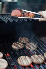 Juicy burger patties sizzling on a charcoal grill with glowing embers below. A hand uses tongs to toast burger buns on the upper rack. Backyard barbecue cooking in progress.