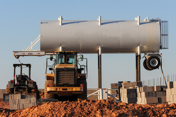 A raised water tanker stands over construction equipment and stacked concrete blocks at a worksite, supporting earthmoving operations and dust control in early-stage infrastructure development