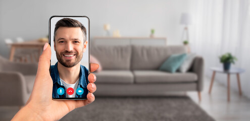 Naklejka premium A person is holding a smartphone displaying a smiling individual during a video call. The setting is a cozy living room with a couch, demonstrating modern telecommunication.