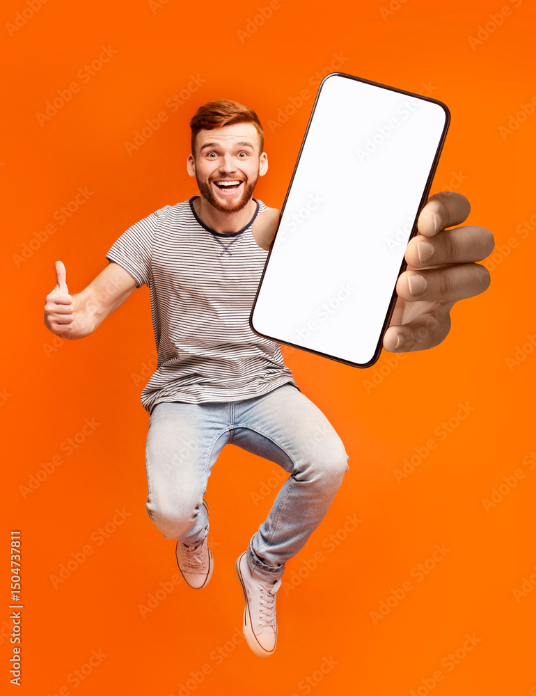 Poster Happy redhead man jumping up in air, showing thumbs up gesture, demonstrating smartphone with empty screen, recommending new mobile application on orange studio background