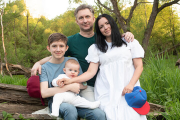 Fototapeta premium Family posing for photo in park. Two children with parents. Teenage boy, baby, mom and dad sitting on log outdoors in summer.