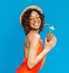 Happy black girl in swimsuit enjoying exotic summer coconut cocktail and smiling at camera, posing over blue background in studio, copy space