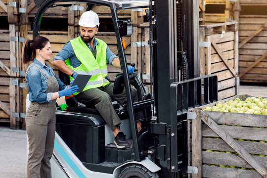 Production of juices and purees, work in warehouse, modern devices for managing loading. Happy young woman show tablet to man in hard hat in forklift with green apples, on wooden boxes background - Powered by Adobe