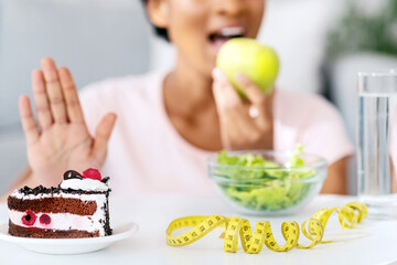 Unrecognizable black woman eating fresh apple and refusing unhealthy cake, blank space. Cropped view of young lady enjoying her fruit, choosing balanced healthy nutrition over junk food