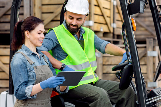 Smart warehouse management system. Millennial bearded man in helmet in forklift truck, discussing order with smiling woman manager in protective gloves with tablet, a lot of wooden boxes background - Powered by Adobe