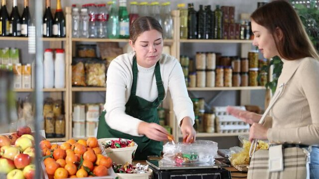Saleswoman in an apron offers chocolates in a basket behind the counter. Buyer chooses the taste of chocolate candies with the help of a seller in a confectionery store
