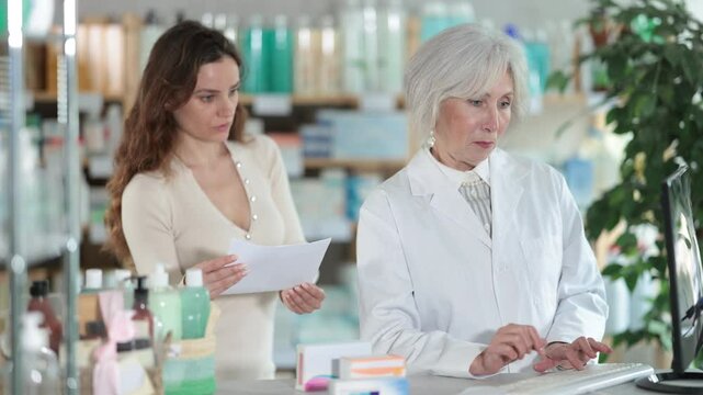 Senior woman employee in labcoat works in pharmacy using computer and collaborate with female client. Professional comment doctors prescription, recommend drug for treatment regiment