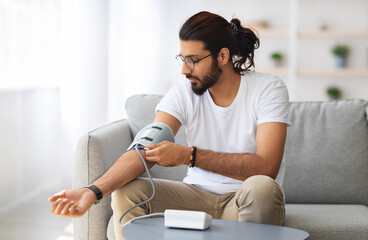 Young indian man sitting on couch at home, checking blood pressure. Middle-eastern guy in white t-shirt using modern tonometer, experiencing hypertension caused by stress, copy space