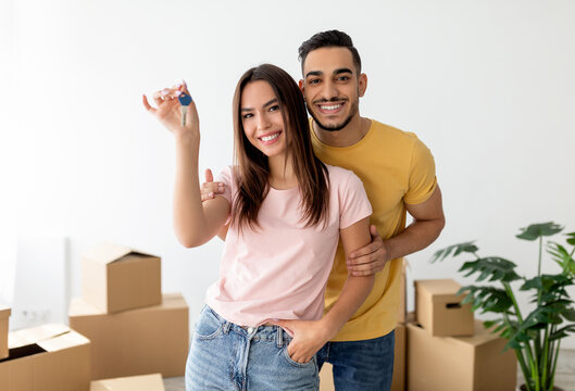 Happy multiracial couple showing key, moving to new apartment together. Affectionate young family posing in their house on relocation day, surrounded by carton boxes - Powered by Adobe