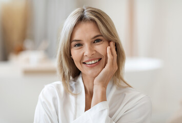 Portrait of smiling beautiful middle aged woman touching her cheek, demonstrating her smooth face skin after using anti-aging cosmetics in the morning, bathroom interior, closeup, copy space