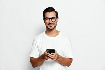 Fototapeta premium Smiling man wearing glasses holds smartphone in hand, staring at camera in casual white t-shirt against plain solid background. Lifestyle concept.