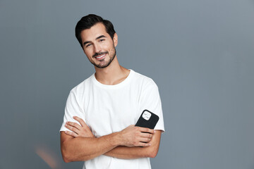 Smiling man with crossed arms holds smartphone while standing against a plain gray background....