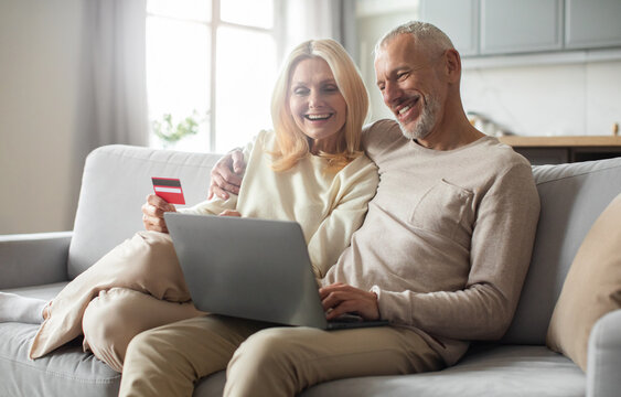 A joyful senior couple sits together on a sofa, engaging in online shopping. They share smiles, with a credit card in hand, enjoying the comfort of their home in the morning light.