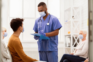 Obraz premium Coronavirus Medical Background. Black Doctor Talking With Sick Asian Patient Guy About Covid-19 Disease Symptoms In Hospital Waiting-Room. Corona Virus Pandemic Concept. Free Space