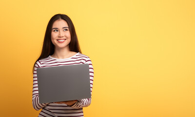 A cheerful young woman dressed in a striped shirt is holding a laptop. She stands against a vibrant...