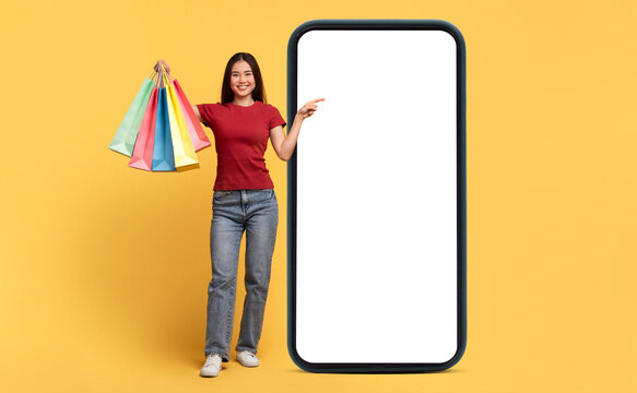 A young woman stands against a bright yellow background, happily holding colorful shopping bags. She points at a large blank phone screen, suggesting an engaging mobile experience.