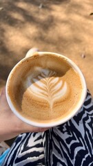 Realistic vertical image of the top view of someone holding a cappuccino with latte art made with fresh milk and drinking her coffee outdoor at daytime in a park in Tokyo Japan.