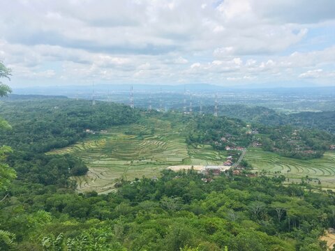 A vast panoramic view from a hilltop, showcasing rolling green hills, dense forests, agricultural fields, and distant villages under a bright, partly cloudy sky.
