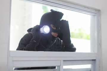 Thief Wearing Black Mask Peeking into House Through Window or Glass Door Using Flashlight