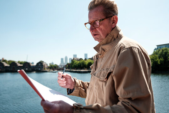 Senior engineer examining blueprint by the river with canary wharf in background