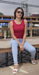 Fototapeta premium Stylish young Asian woman in a red top and blue jeans sits on a wooden bench on outdoor stands, showcasing a modern, casual look with platform sandals
