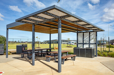 Contemporary outdoor barbecue shelter with picnic benches and stainless steel BBQ facilities in a well-maintained suburban park in Australia, offering shade and comfort for public gatherings