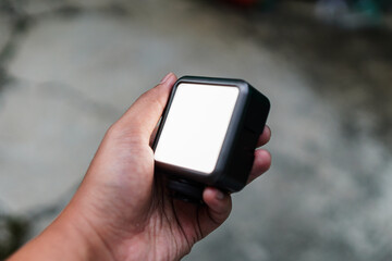 A hand holds a compact, illuminated LED light panel against a blurred neutral background. The black-edged panel provides bright, even illumination for photography and video projects.