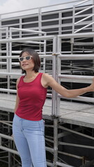 Fototapeta premium Pretty Asian woman stands on outdoor metal stands, showcasing modern casual fashion with a bold red sleeveless top and light blue jeans
