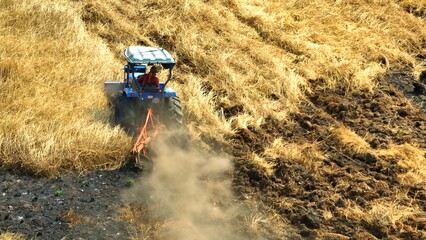 A bird's eye view captures the rustic elegance of a tractor carving through arid soil, leaving a billowing cloud of dust in its wake, amidst the warm hues of straw-covered land.
