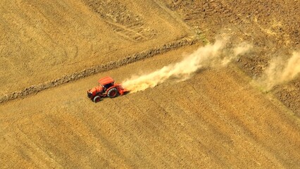 Across the sun-scorched land, a tractor hauls a cultivator, leaving a trail of swirling dust in its...