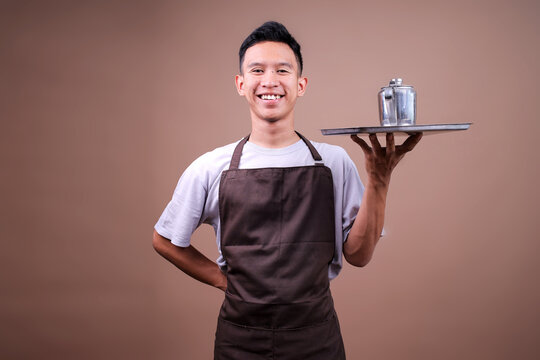 Cheerful Young Barista Guy Wearing Brown Apron Holding Serving Tray With Coffee Or Tea Pot