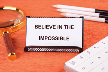 Motivational quote. BELIEVE IN THE IMPOSSIBLE text on the card next to a magnifying glass, pencils...