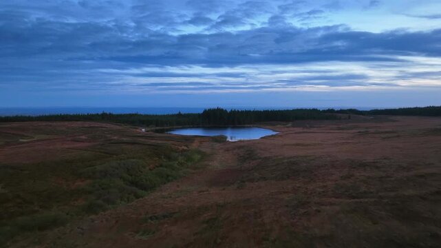 Bragan, County Monaghan, Ireland, September 2022. Drone descends along hills over Lake Menish with textured clouds and dense forest reflecting in waters, panoramic aerial establishing at blue hour.