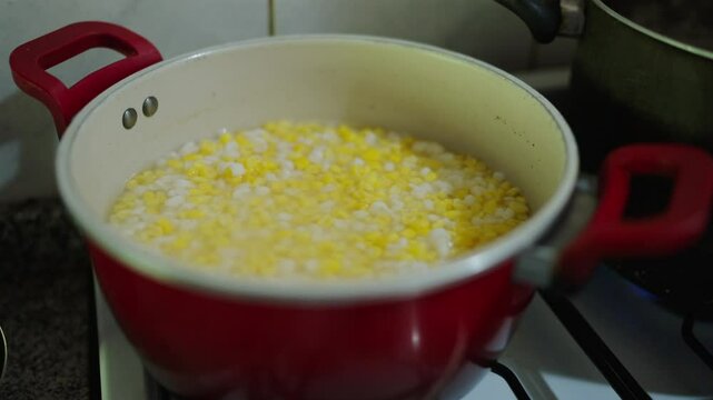 Bright yellow and white corn kernels simmer in a red enamel pot on a stove, bubbles rising as the grains soften in water, evoking the start of a creamy porridge or chowder in a cozy home kitchen scene