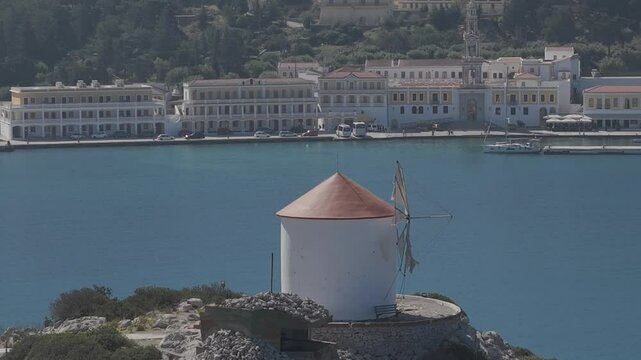 Stunning aerial perspective of the historic Panormitis Monastery on Symi. The footage showcases traditional Byzantine architecture in the background behind a windmill. 3x telephoto lens. D-Log M