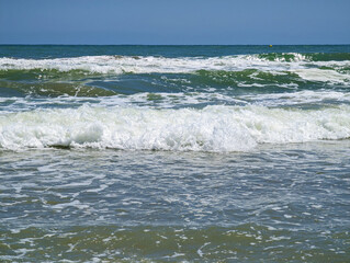 Foamy waves of Mediterranean beach of Mareny de Barraquetes on sunny summer day in Valencia, Spain. Blue and green sea water, white choppy foam, clear sky, paradisiacal seashore, coastal nature. 