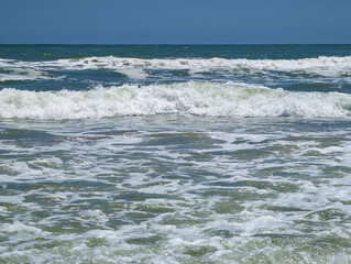 Foamy waves of Mediterranean beach of Mareny de Barraquetes on sunny summer day in Valencia, Spain. Blue and green sea water, white choppy foam, clear sky, paradisiacal seashore, coastal nature.