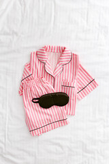 Pink white Pajama Set with black silk sleep mask on white sheet. Top view layout fashion woman home sleepwear from natural cotton on bedclothes. Cozy pyjamas of shirt, shorts for comfort dreams