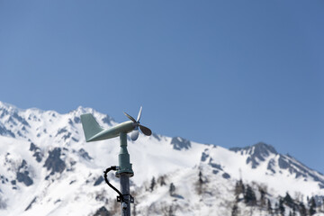 春の青空と雪山を背景に、黒部平パノラマテラスに設置された風向風速計（気象観測装置）が立つ様子。
