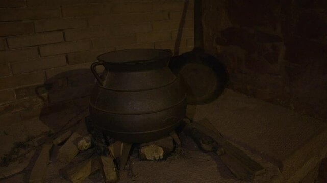 A large cast iron cauldron sits on a stone hearth above firewood in a dimly lit, rustic kitchen or medieval fireplace, evoking scenes of historical cooking or potion brewing.